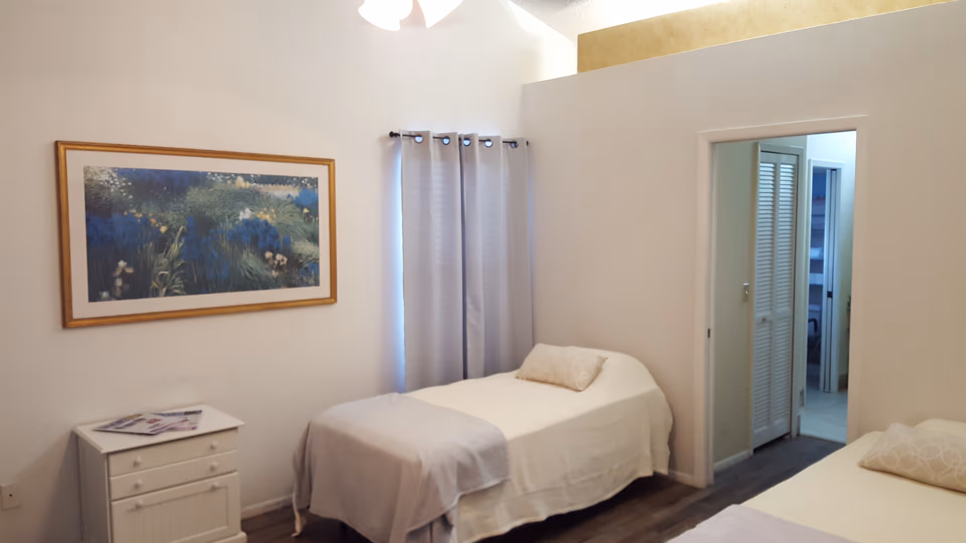 A simple bedroom with two single beds covered in white linens and light-colored pillows. There is a small white nightstand with drawers between the beds, holding some magazines. A framed painting of a nature scene hangs on the wall above the nightstand. A window with light gray curtains is on the wall behind one of the beds. The room has light-colored walls and dark wood flooring. A doorway leads to another room with visible closet doors.