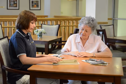 A caregiver and an elderly woman sitting at a wooden table in a common area, looking at photographs together. The caregiver is wearing a navy blue polo shirt and the elderly woman is dressed in a light pink blouse. The background shows more tables and chairs in a well-lit room.