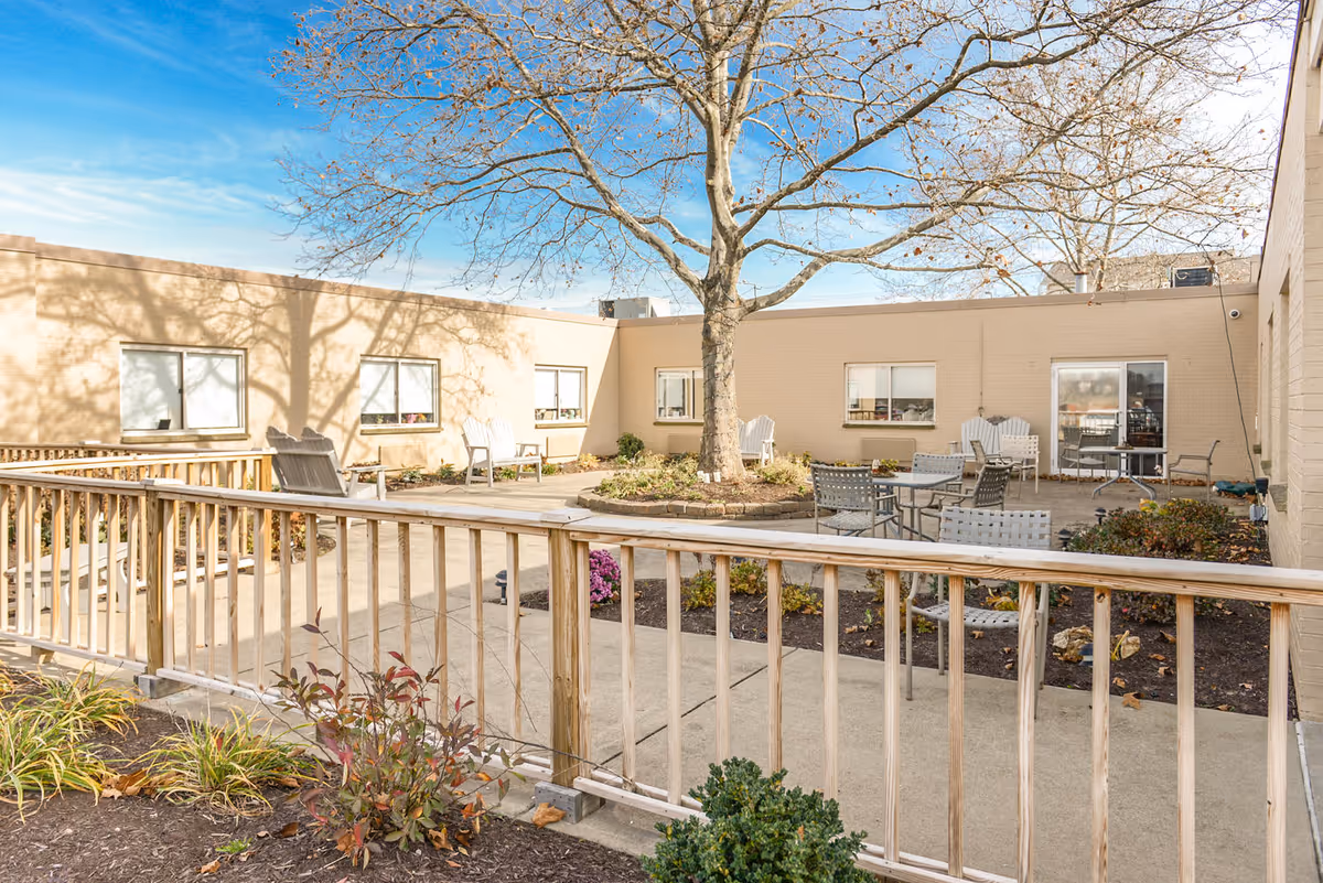 Outdoor courtyard area at Morgantown Healthcare Center featuring a large tree in the center surrounded by a circular garden bed. There are several chairs and tables arranged on the concrete patio, with beige building walls and windows enclosing the space. A wooden railing runs along the foreground with some plants and shrubs in the garden beds.