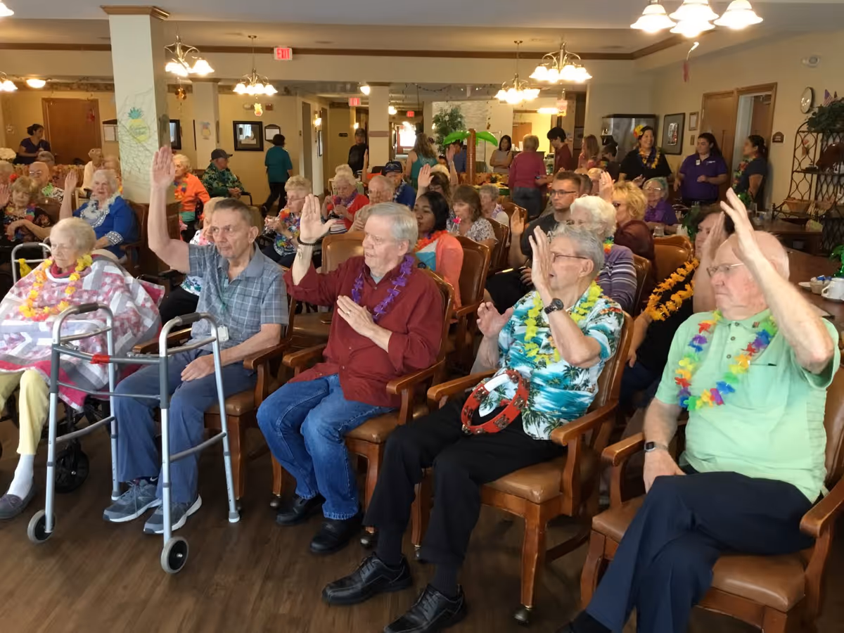 A group of elderly people seated in a community room, many wearing colorful leis and raising their hands. The room is well-lit with wooden floors and decorated with plants and wall art. Several staff members and other residents are visible in the background.