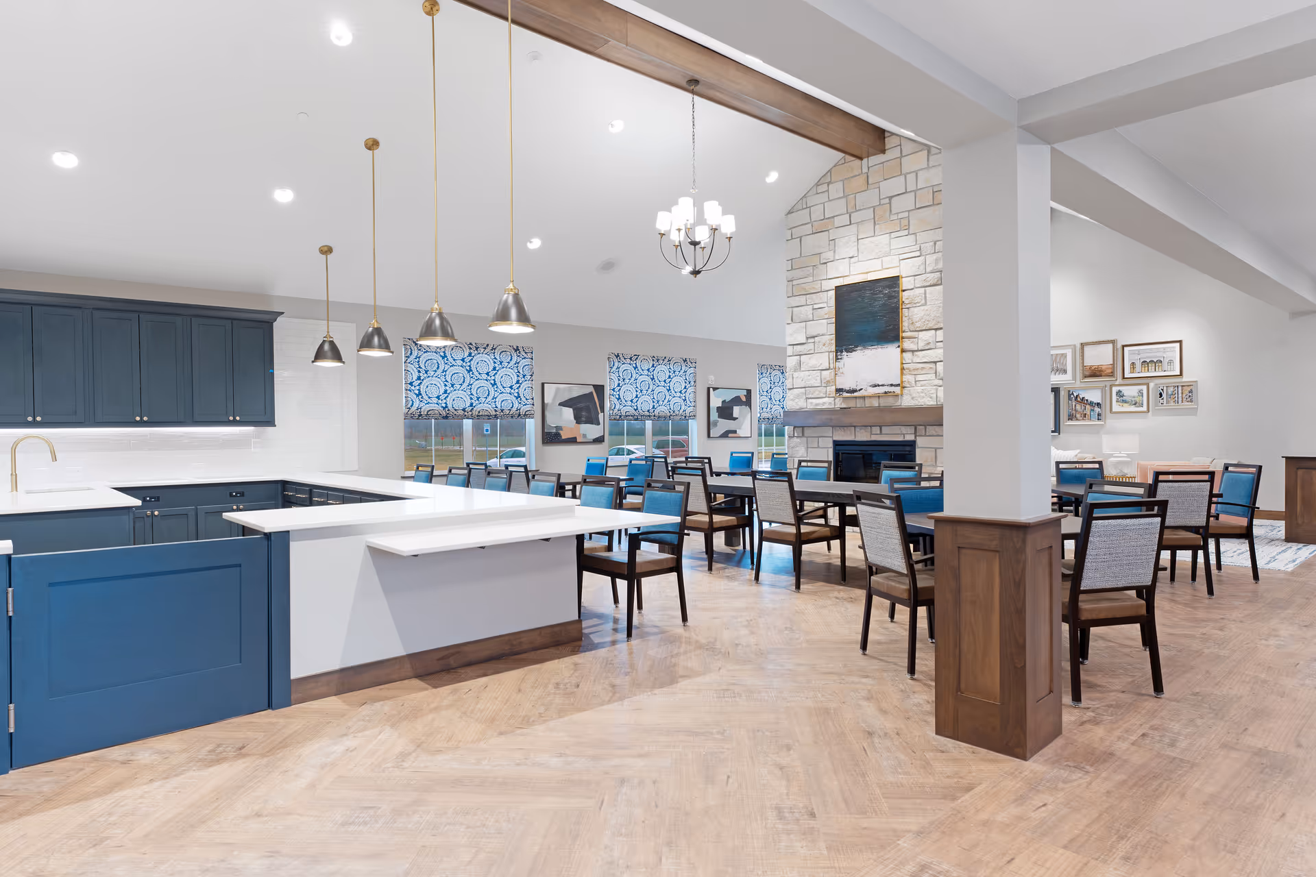 A spacious senior living dining area with multiple tables and chairs arranged neatly. The room features a modern kitchen with blue cabinetry, white countertops, and pendant lighting. Large windows with patterned blue and white shades allow natural light to fill the space. A stone fireplace with a painting above it is visible in the background, and the floor is covered with light wood flooring.