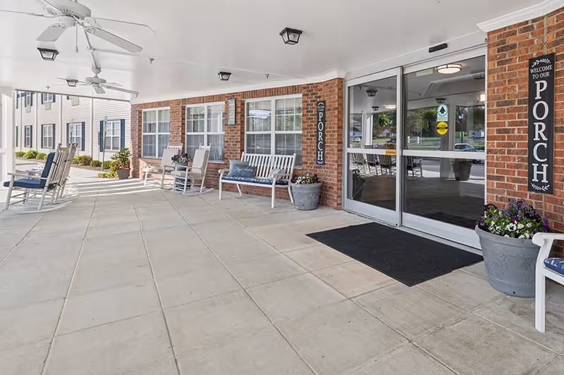 Covered porch area at Home Place of Burlington with white rocking chairs and benches, ceiling fans, potted plants, and sliding glass entrance doors. The porch has a brick wall with windows and signs that say 'Welcome to our Porch'.