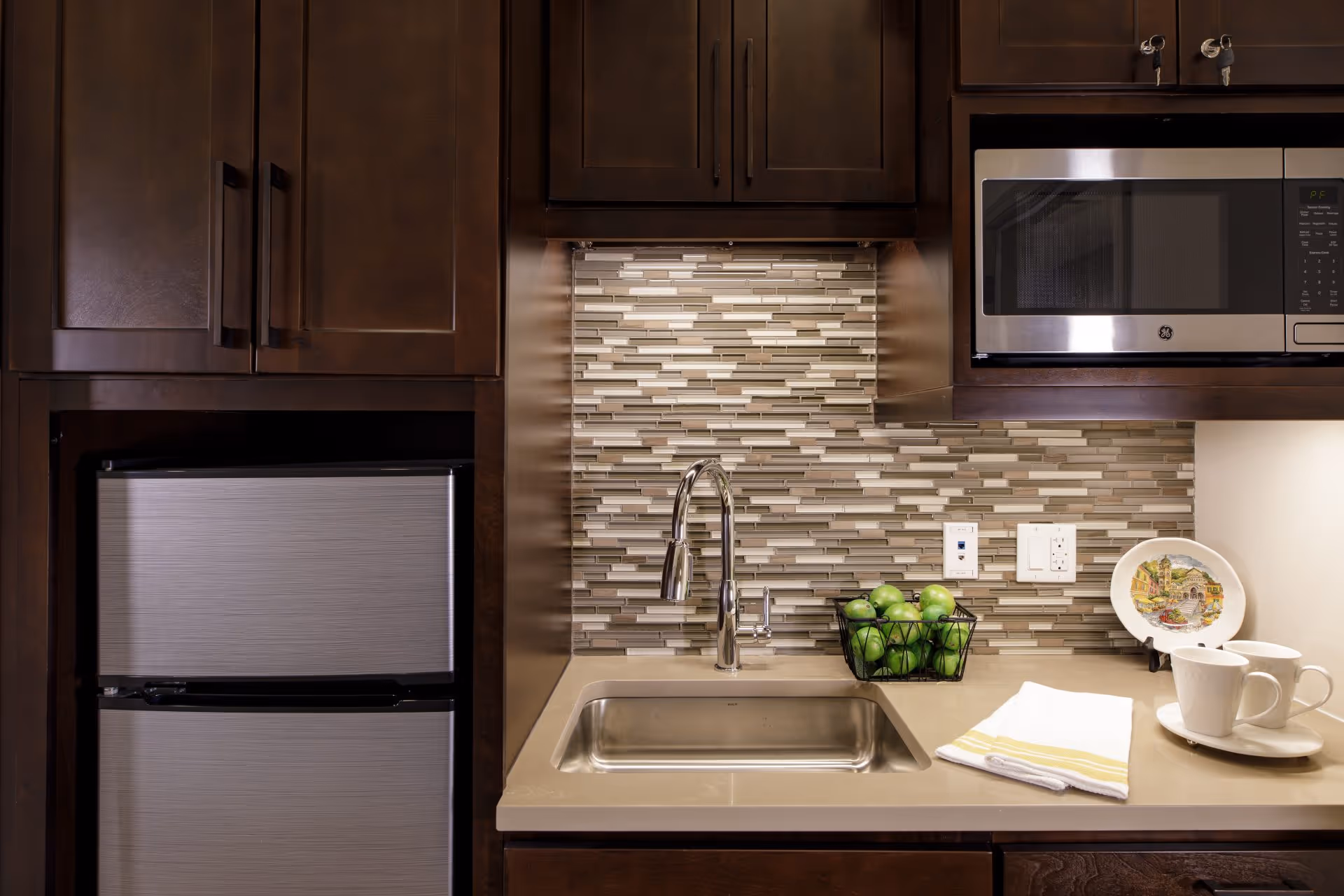 A modern kitchen area with dark wooden cabinets, a stainless steel mini refrigerator, a built-in microwave, a sink with a chrome faucet, a basket of green apples, a decorative plate, and two white cups on saucers with a folded towel on the countertop.