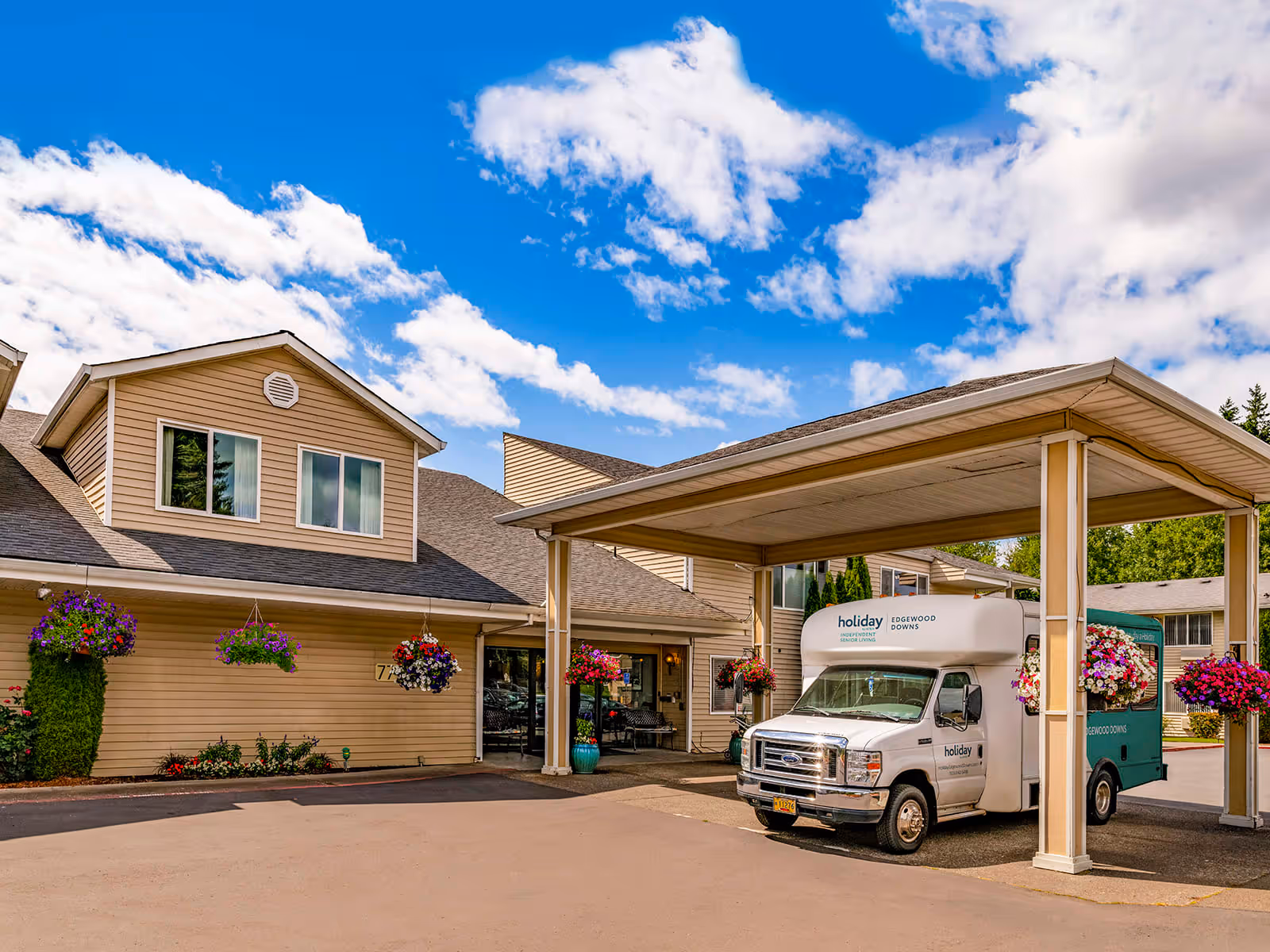 Exterior view of Holiday Edgewood Downs facility showing the entrance with a covered drop-off area. A white and green shuttle van with the facility's name is parked under the canopy. The building is beige with hanging flower baskets and a clear blue sky with some clouds above.