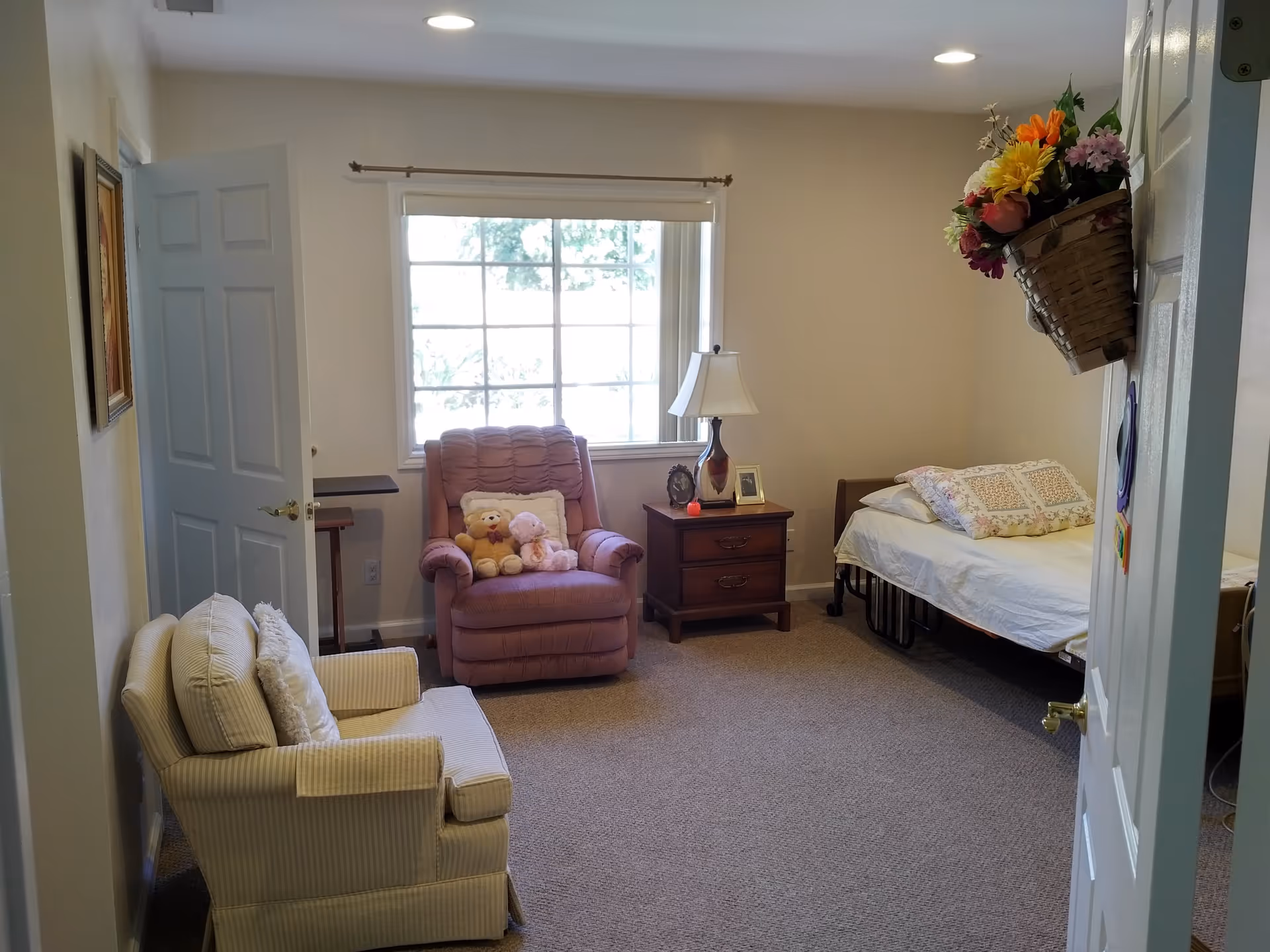 A cozy assisted living room with a single bed on the right side, a wooden nightstand with a lamp and framed photos, a pink recliner chair with two stuffed animals, and a beige armchair with a pillow. A large window with a white blind is centered on the back wall, and a basket with colorful flowers hangs on the right wall near the door.