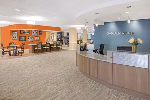 Spacious senior living lobby with a curved reception desk labeled "GARDEN RIDGE" and a dining/seating area in the background.