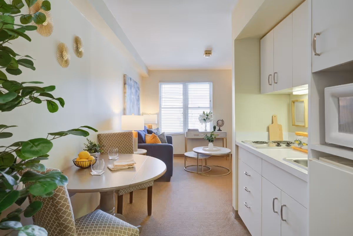 A bright and cozy studio apartment featuring a small kitchen area with white cabinets, a microwave, and a stove on the right. On the left, there is a round dining table with two patterned chairs and a bowl of lemons. In the background, a living area with a blue sofa, colorful cushions, a side table with a lamp, a coffee table with a plant, and a window with white blinds letting in natural light. Decorative wall art and a green plant add warmth to the space.