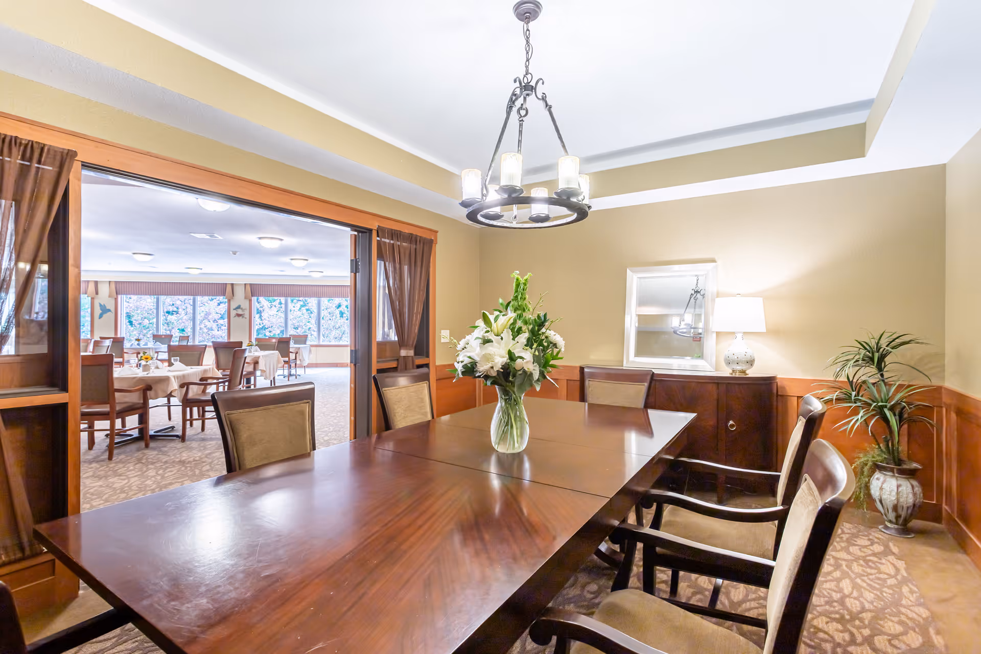 A well-lit dining room with a large wooden table surrounded by six chairs. A vase with white flowers is placed in the center of the table. The room has beige walls, a decorative ceiling light fixture, a sideboard with a lamp and a mirror, and a potted plant in the corner. Through an open doorway with wooden trim and sheer brown curtains, another dining area with multiple tables and chairs is visible, featuring large windows letting in natural light.