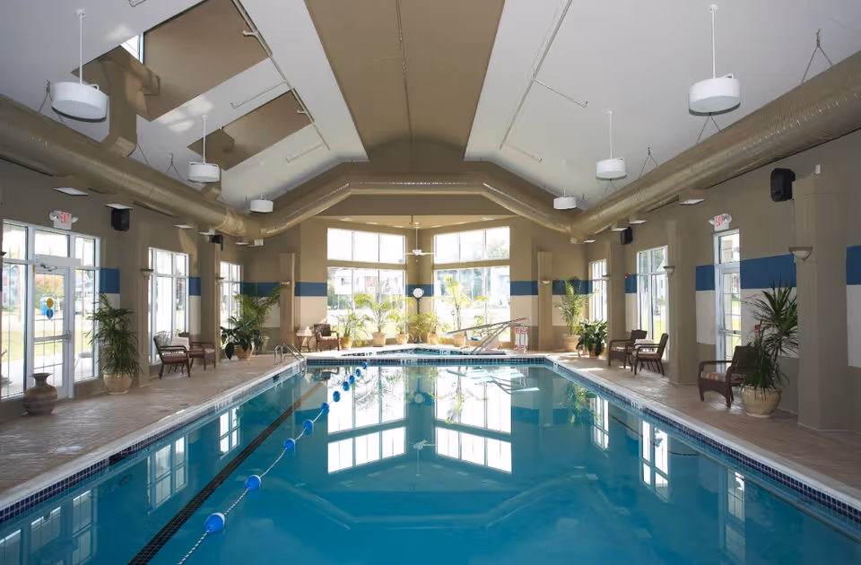 Indoor swimming pool with clear blue water, surrounded by large windows letting in natural light. The pool area has several potted plants and chairs along the sides. The ceiling features exposed ductwork and hanging light fixtures.