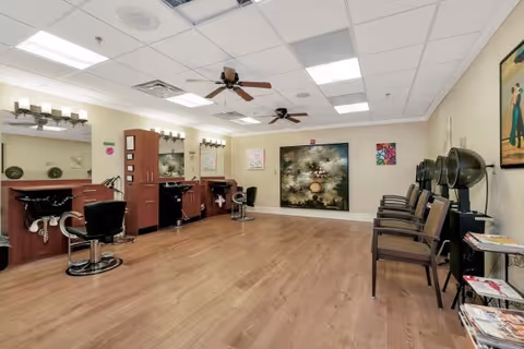 Bright salon room with styling stations, shampoo sinks, salon chairs and a row of waiting chairs under ceiling fans.