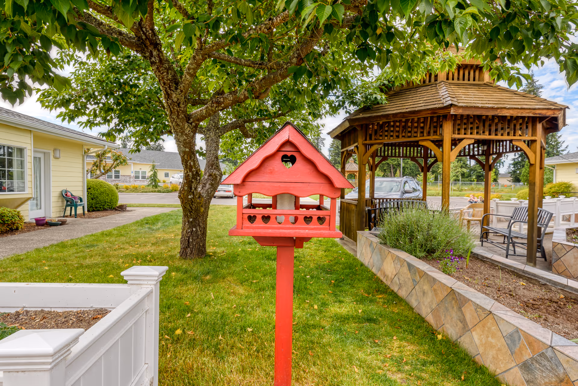 A red birdhouse on a post stands in a grassy area with a large tree behind it. To the right, there is a wooden gazebo with seating and a tiled planter box with greenery. On the left, there is a yellow building with windows and a pathway leading to it. The sky is partly cloudy.