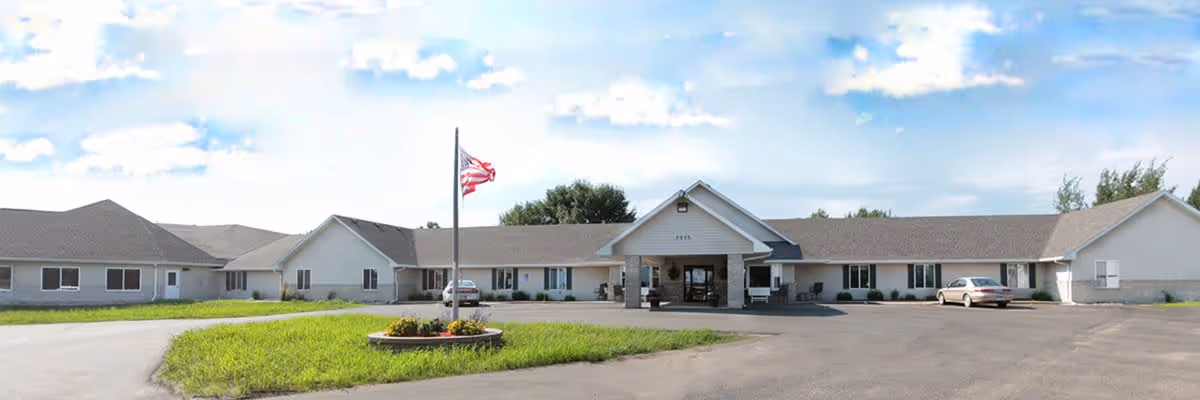 Exterior view of a single-story senior living facility building with a peaked entrance, an American flag on a flagpole in front, a circular driveway, and two parked cars. The sky is partly cloudy.