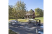 Outdoor patio area with picnic tables, chairs, an umbrella and a small gazebo on a grassy lawn surrounded by trees.
