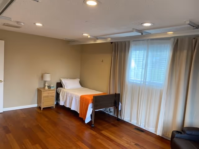A simple bedroom with a single bed against the wall, covered with white bedding and an orange throw blanket. Next to the bed is a wooden nightstand with a lamp and a small item on it. There is a window with sheer white curtains letting in natural light, and the floor is wooden. The room has beige walls and a white ceiling with recessed lighting.