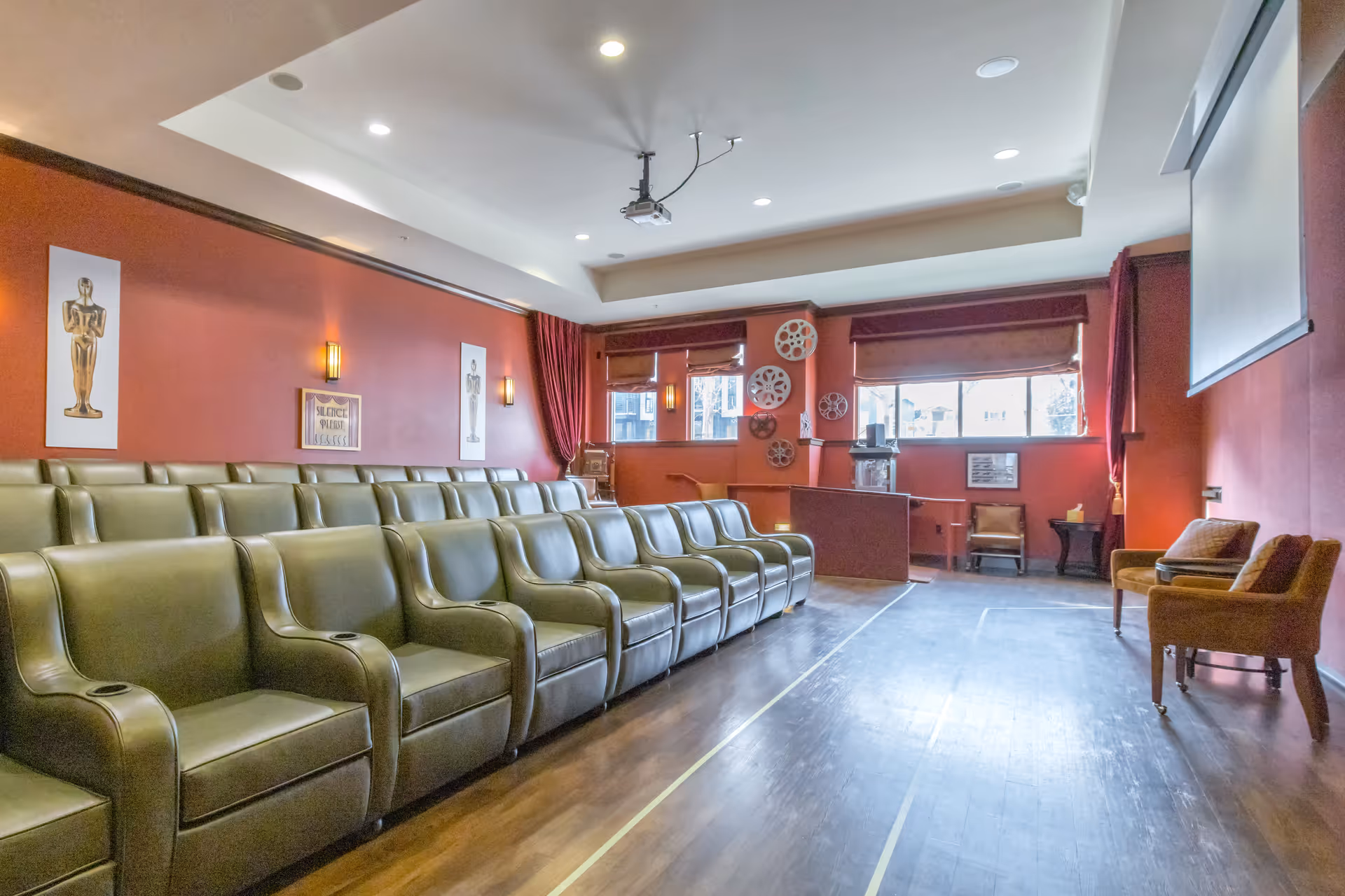 A small theater room with two rows of green leather armchairs facing a large projector screen. The walls are painted red and decorated with movie-themed artwork, including golden statuettes and film reels. There are windows with brown blinds and red curtains, and the floor is wooden.