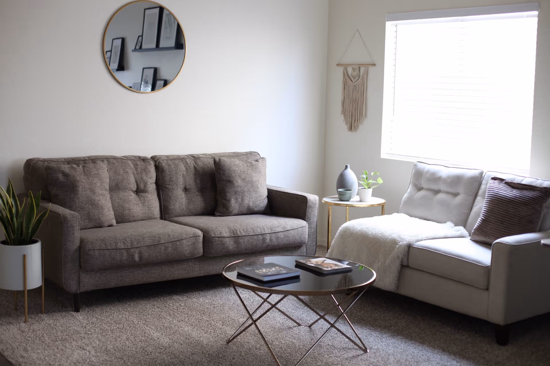 A cozy living room with two sofas, one brown and one light gray, arranged around a round glass coffee table with books on it. There is a small round side table with decorative items and a plant next to the light gray sofa. A round mirror and a macrame wall hanging decorate the white walls, and a window with blinds lets in natural light.