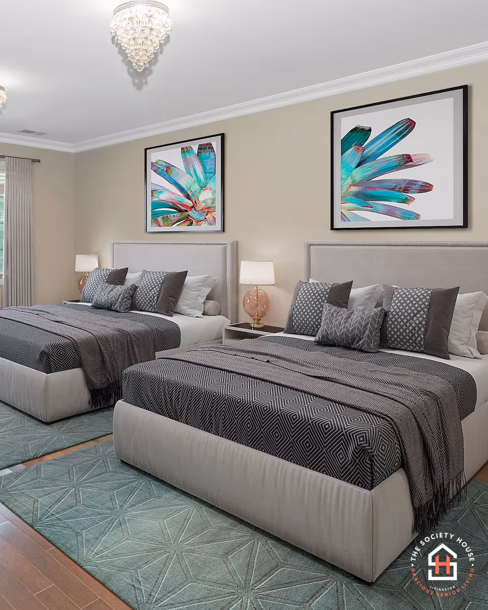 A bedroom with two large beds featuring beige upholstered headboards, gray and black patterned bedding, and multiple decorative pillows. Each bed has a nightstand with a lamp that has a pink glass base. Above each bed hangs a colorful abstract painting with teal and purple hues. The room has beige walls, a light blue patterned rug, wooden flooring, and a window with beige curtains.