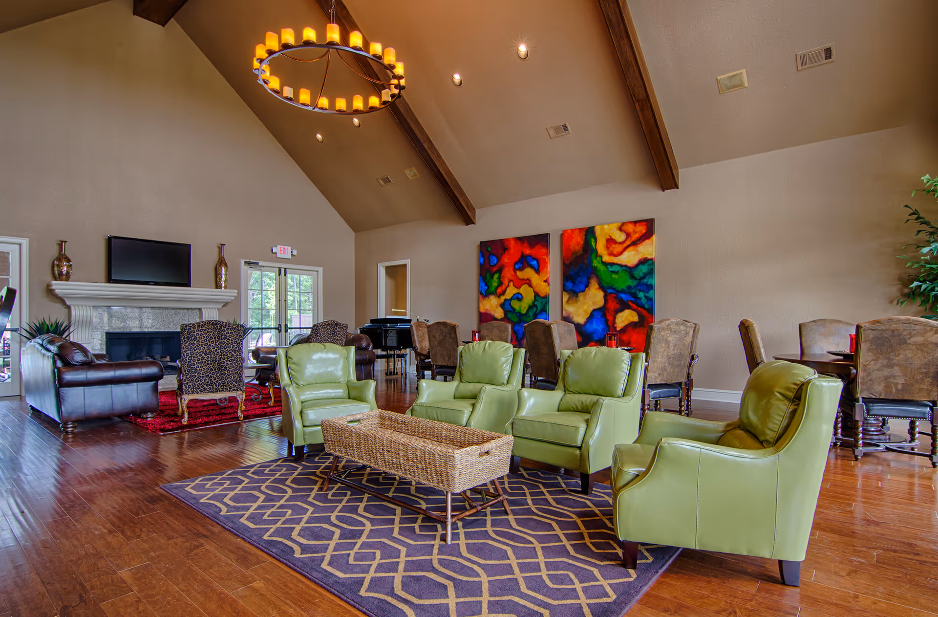 A spacious living room with high vaulted ceilings and exposed wooden beams. The room features a seating area with four green armchairs arranged around a wicker coffee table on a patterned rug. In the background, there is a fireplace with a TV mounted above it, two patterned armchairs, and a red rug. There are also several dining tables with chairs and colorful abstract artwork on the wall.