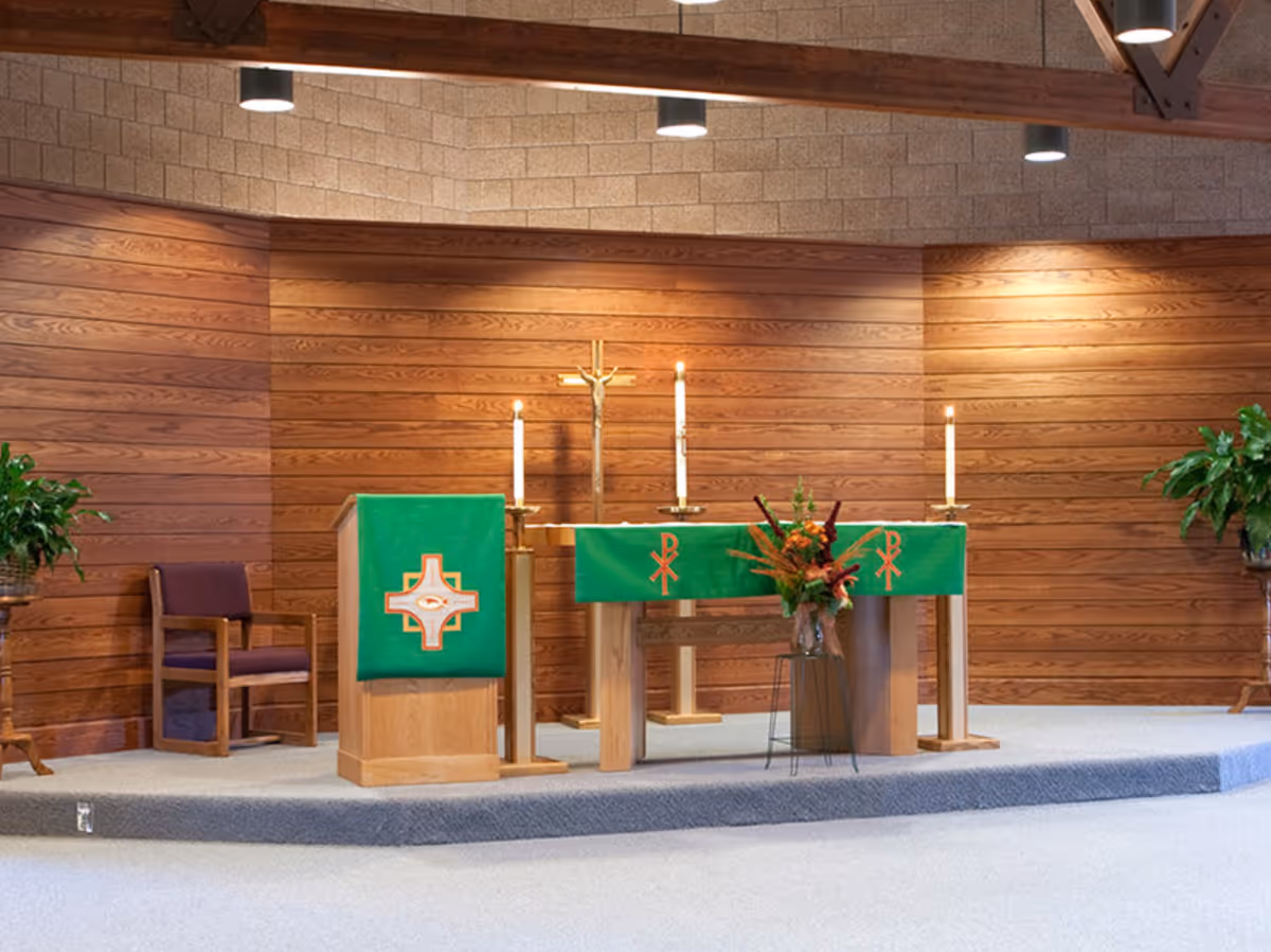 A wood-paneled chapel interior showing a raised altar draped in green cloths with candles, a cross, flowers, and seating.