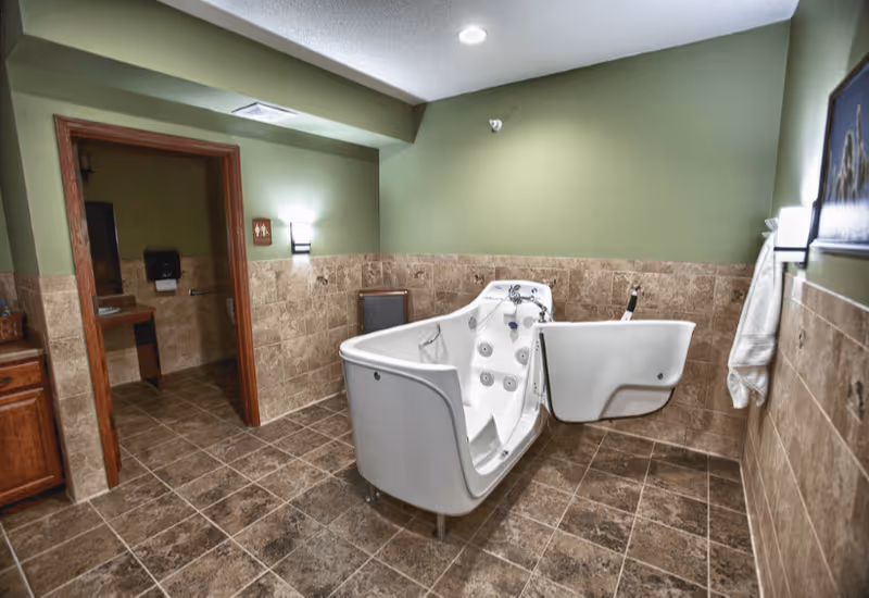 A spacious bathroom with brown tiled floors and walls halfway up, painted green above. The room features a white walk-in bathtub with an open door, designed for accessibility. There is a doorway leading to another room with a sink and a paper towel dispenser visible. Wall-mounted lights provide illumination, and a towel hangs on a hook on the right wall.