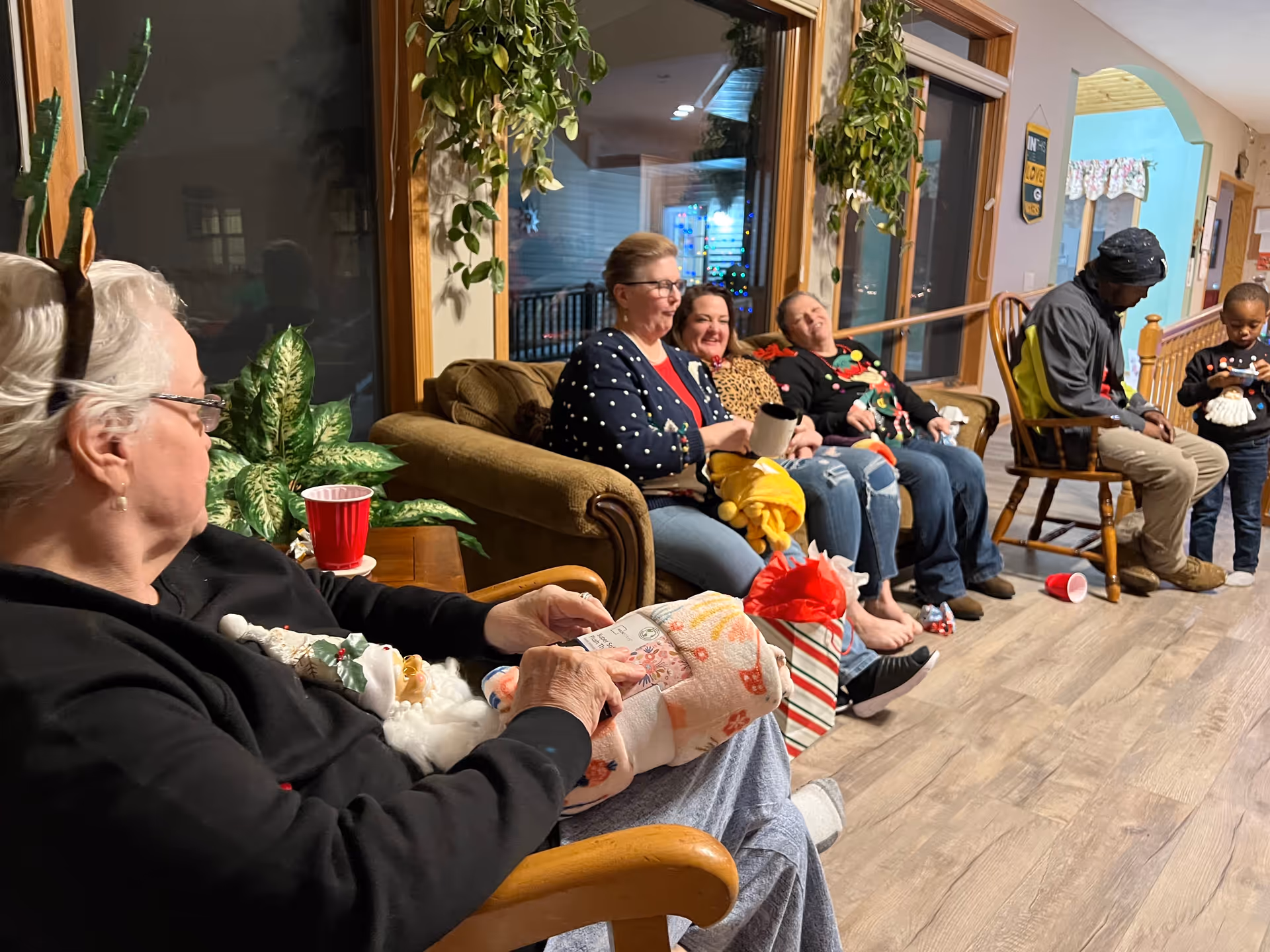 Several people sitting on couches and chairs in a communal living room area, chatting and holding gifts and drinks.