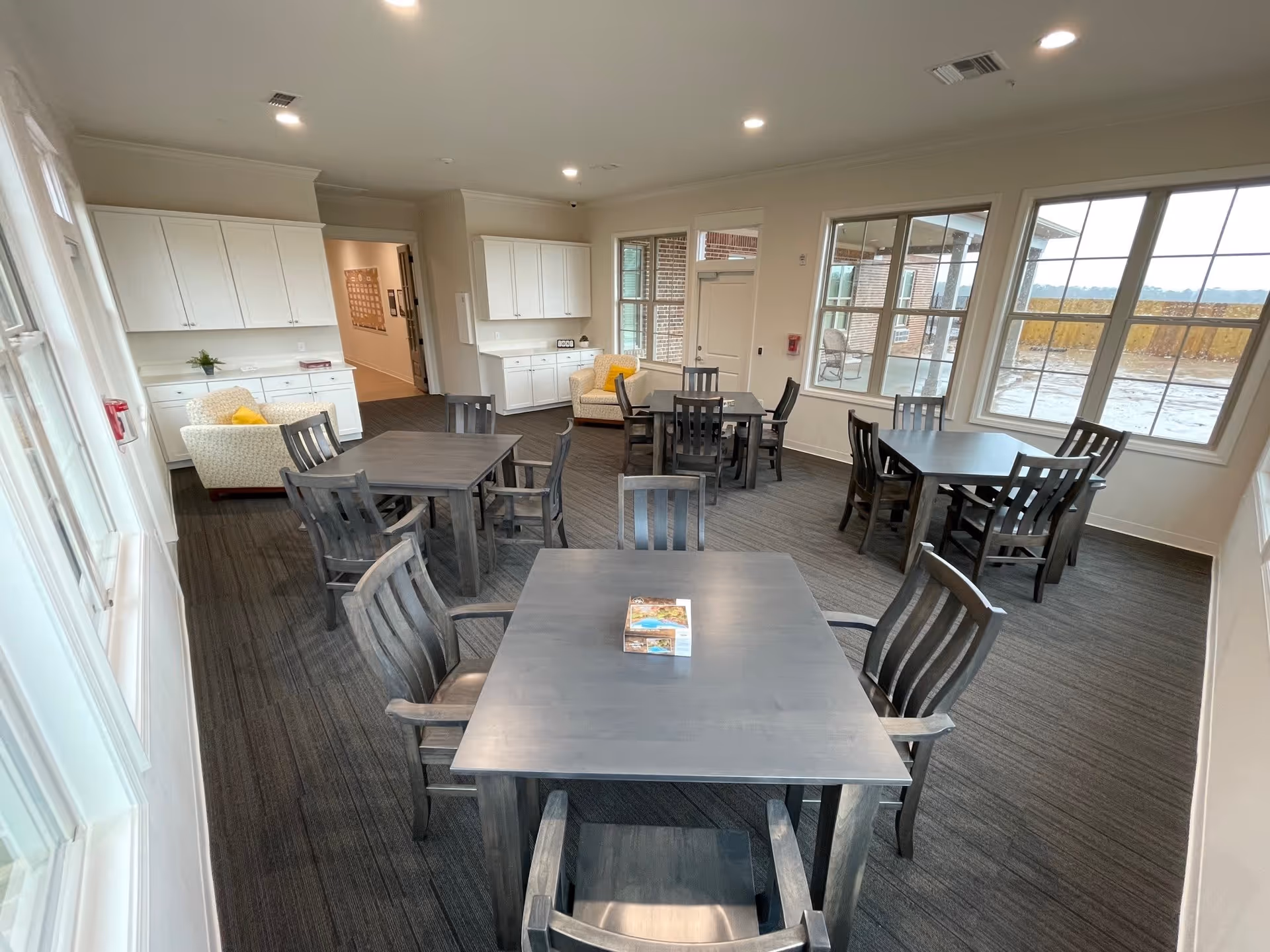 A bright and spacious dining area with several square wooden tables and matching chairs arranged neatly on a carpeted floor. Large windows on two walls allow natural light to fill the room and provide a view of an outdoor patio and landscape. The walls are painted white, and there are white cabinets along one wall with two beige armchairs featuring yellow accent pillows placed near the cabinets. The ceiling has recessed lighting.