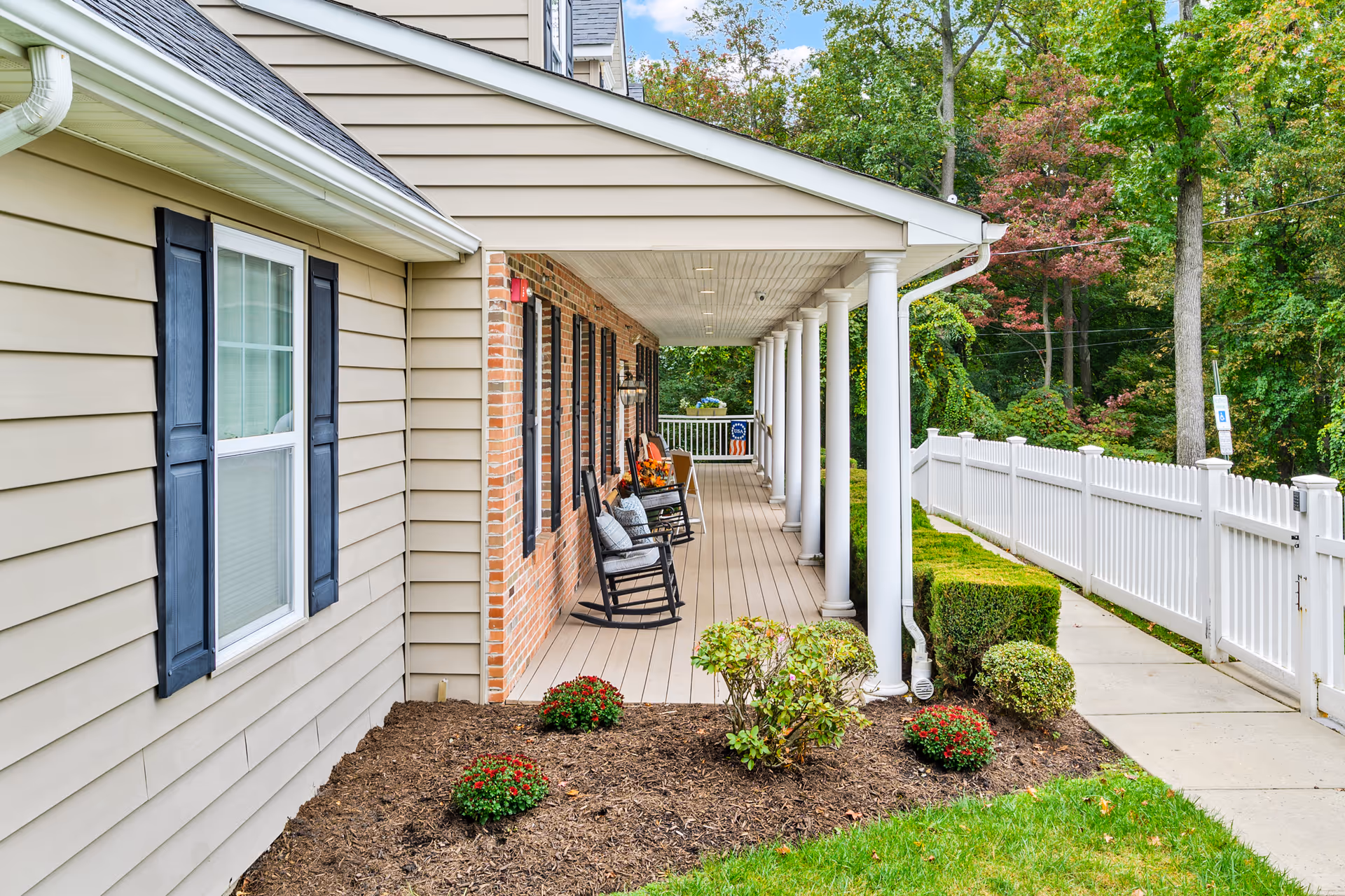 Covered porch area of a residential building with beige siding and brick walls, featuring black rocking chairs with cushions, white columns, and a white picket fence along a sidewalk. There are small bushes and flowers planted in mulch beds, and trees with green and some autumn-colored leaves in the background.