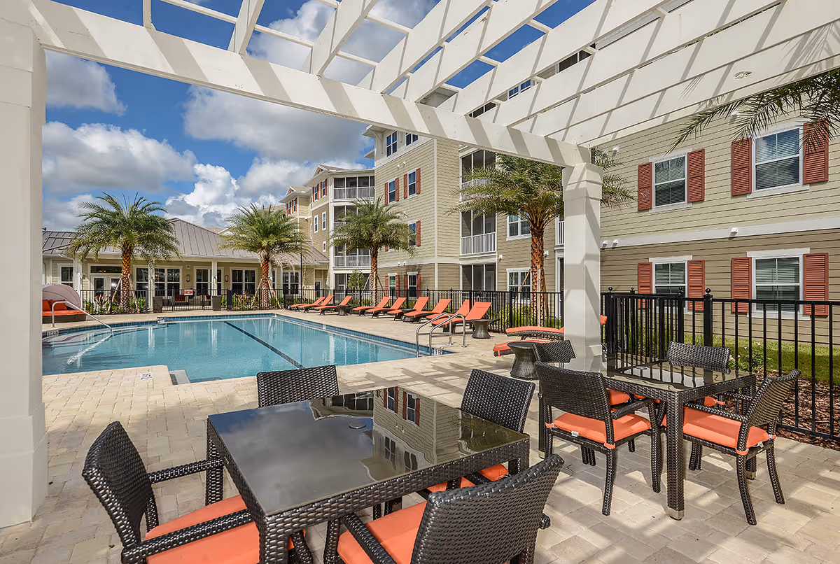 Outdoor pool area at Album Monterey Pointe 55+ Active Adult Apartment Homes featuring a swimming pool surrounded by palm trees, lounge chairs with orange cushions, and patio tables with matching chairs under a white pergola. Beige apartment buildings with red shutters are visible in the background under a partly cloudy sky.