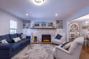Cozy living room with a brick fireplace flanked by built-in shelves, a navy sofa, armchairs, and an area rug with a view into the dining area.