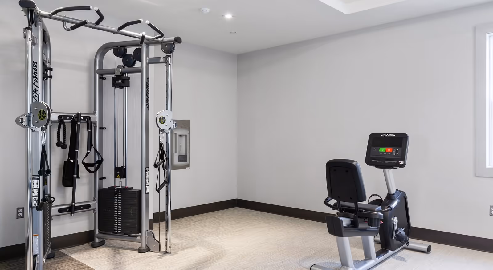 A small fitness room with a Life Fitness multi-functional weight machine on the left and a recumbent exercise bike on the right, set against light gray walls and a window on the right side.