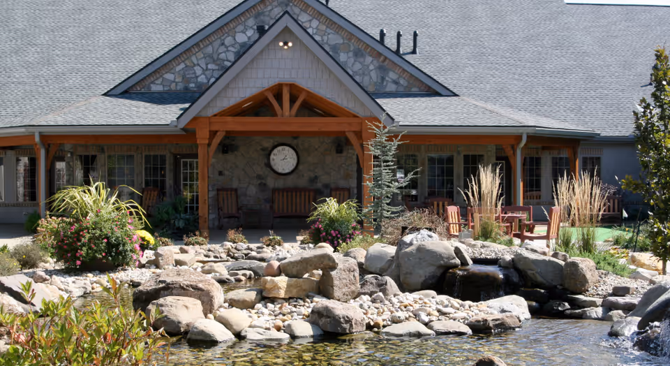 Front entrance of a senior living building with a stone facade and clock above a covered porch, a rock-lined pond and outdoor seating in the foreground.