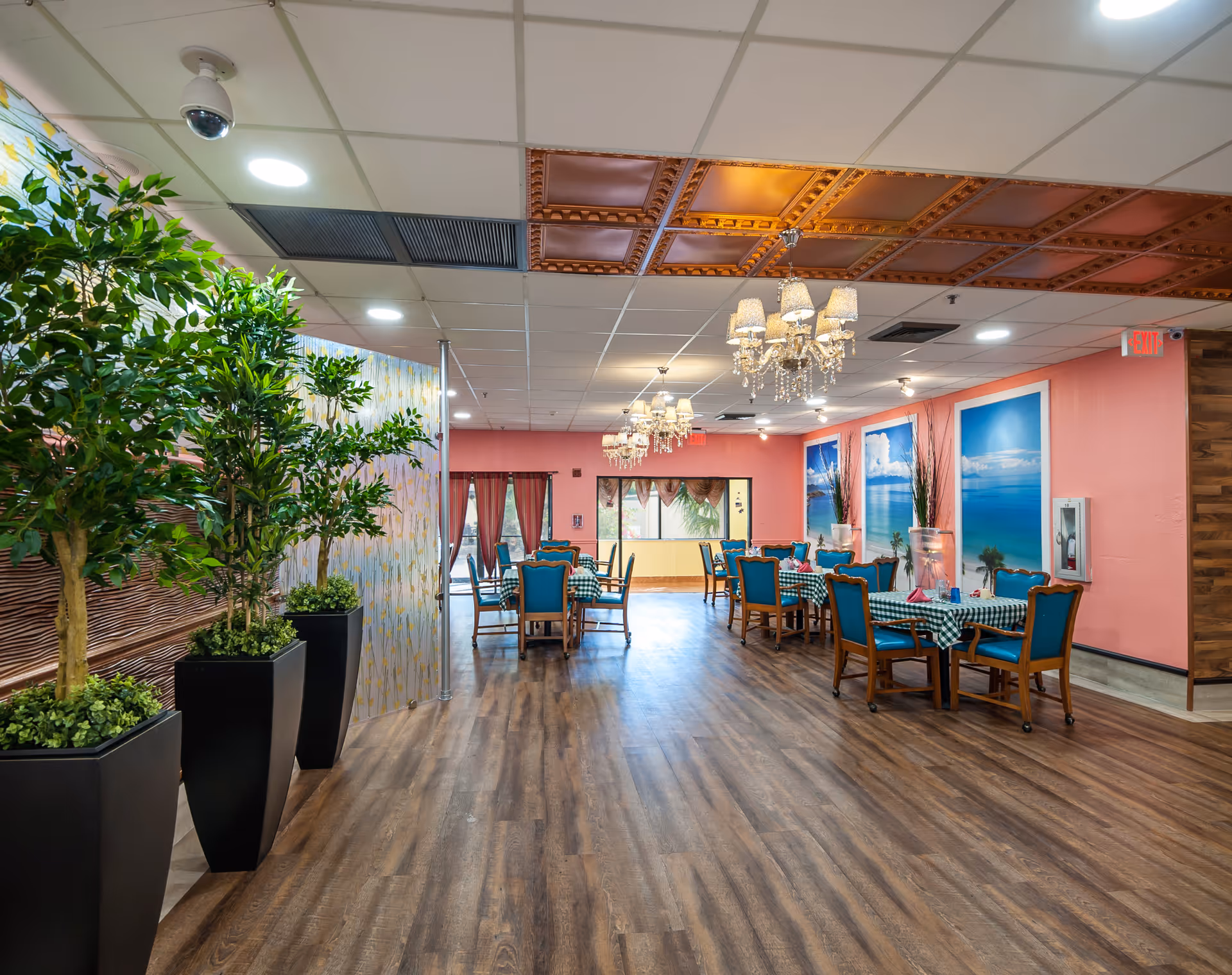 Interior view of a senior living facility dining room with wooden flooring, several tables covered with checkered tablecloths, and blue cushioned chairs. The walls are painted pink and decorated with large framed pictures of beach scenes. There are chandeliers hanging from the ceiling and large potted plants on the left side.