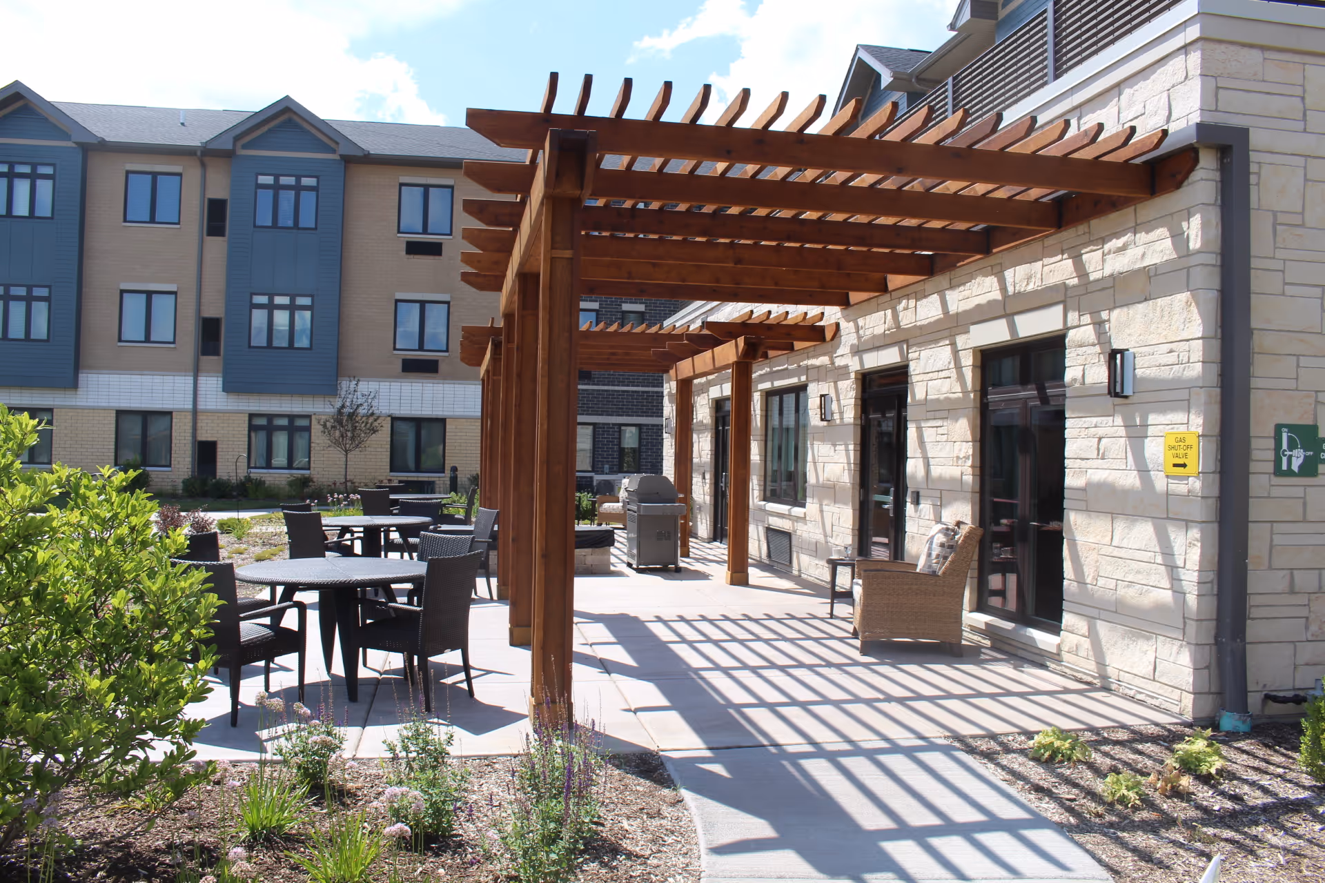 Outdoor patio area at Evergreen Senior Living with wooden pergolas casting shadows on the walkway. There are several black tables and chairs arranged for seating, surrounded by landscaped plants and shrubs. The building exterior features stone walls and multiple windows under a clear blue sky.