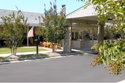 Entrance area of a facility with a covered drop-off zone, surrounded by trees and landscaping, with an American flag visible near the sidewalk.