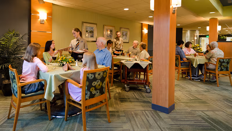 A dining room in The Maplewood Nursing Home with several tables where elderly residents and younger visitors are seated, being served by staff members. The room has green walls, wooden pillars, and patterned carpet flooring, with warm lighting and framed pictures on the walls.
