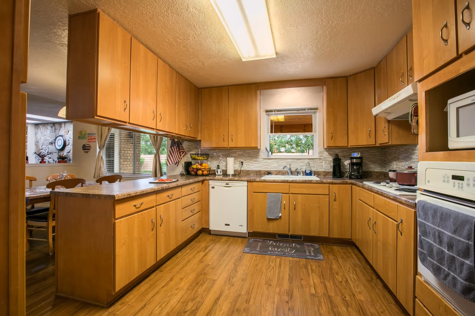A spacious kitchen with wooden cabinets and drawers, a white dishwasher, a white oven, a microwave, a coffee maker, and a sink under a window. The kitchen has a wooden floor and a countertop with a bowl of fruit and some kitchen items. There is a dining area visible through an open space on the left side.