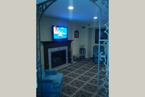 A cozy living room with a patterned carpet, two blue armchairs, a fireplace with a mounted flat-screen TV above it, and a small side table with a decorative item. The room is softly lit with recessed ceiling lights.