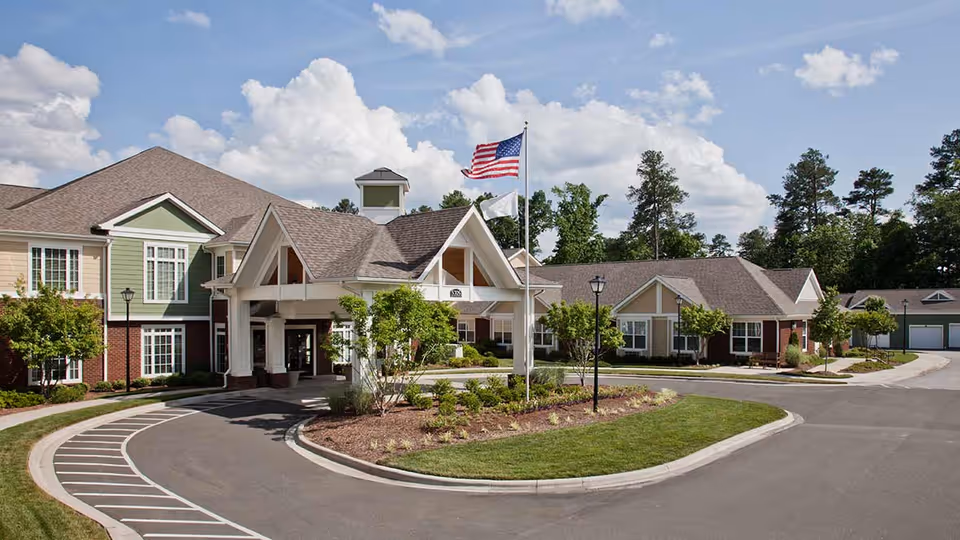 Exterior view of Atria Southpoint Walk senior living facility showing a large building with a covered entrance, landscaped greenery, an American flag, and a clear sky with clouds.