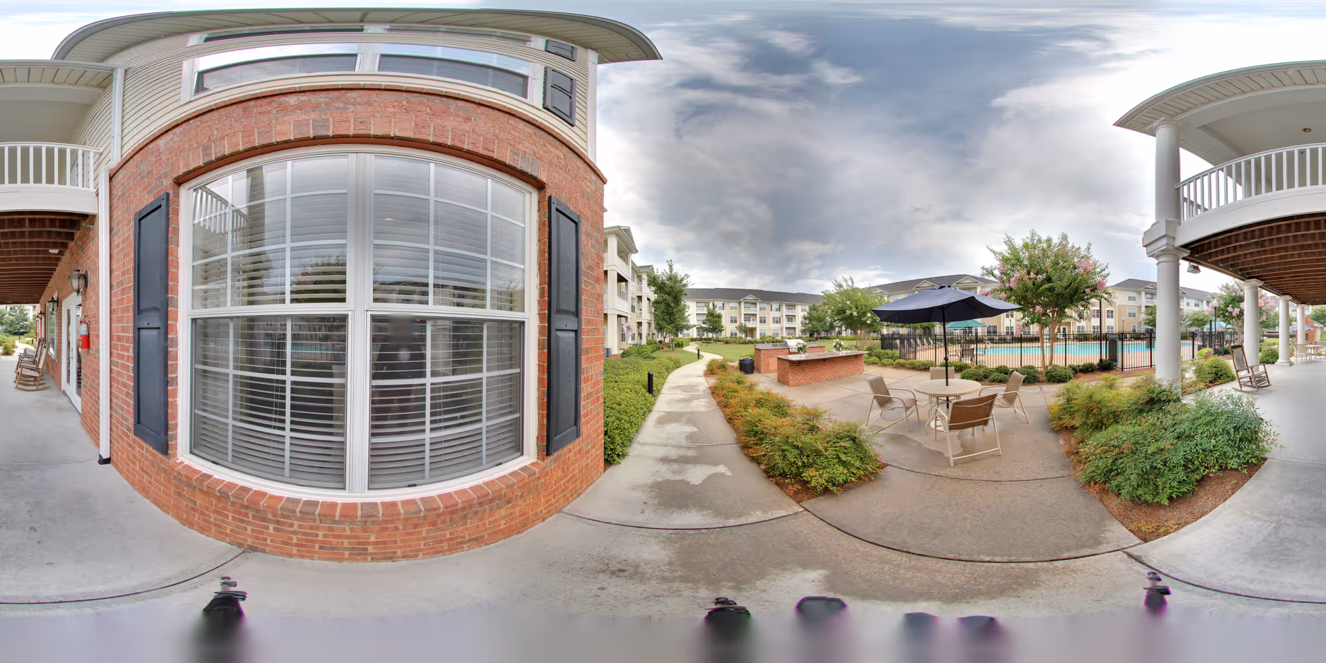 Outdoor patio area at The Palms at Lake Spivey featuring a concrete walkway, round table with chairs and an umbrella, surrounded by bushes and trees. In the background, there is a fenced swimming pool and multi-story residential buildings. The sky is partly cloudy.