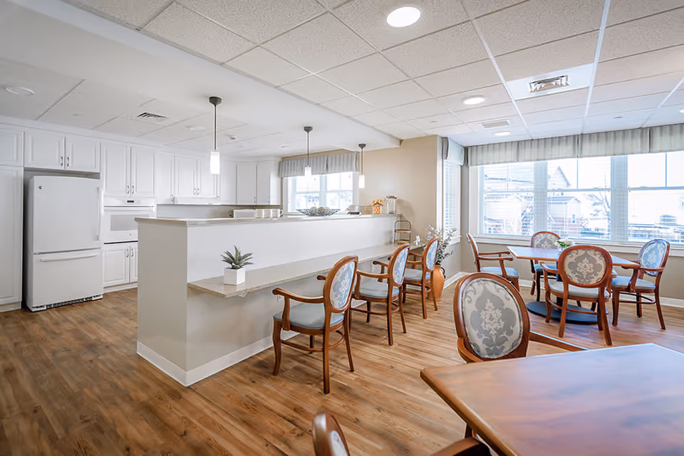 A bright and clean dining area with wooden floors and multiple tables and chairs. The room features a kitchen area with white cabinets, a refrigerator, and a counter with bar stools. Large windows allow natural light to fill the space.