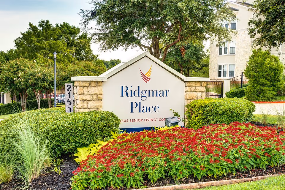 Outdoor view of the entrance sign for Ridgmar Place senior living community, surrounded by well-maintained landscaping including green bushes, red flowers, and trees, with part of the building visible in the background.