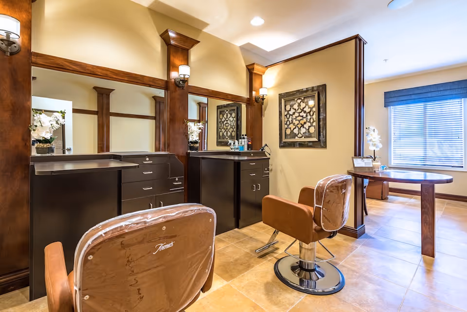Interior view of a senior living facility's salon area with two brown salon chairs facing large mirrors mounted on wooden framed walls. The room has beige tiled floors, warm lighting from wall sconces, and a window with blinds letting in natural light. A decorative framed artwork hangs on the wall, and a small table with a flower arrangement is visible near the window.