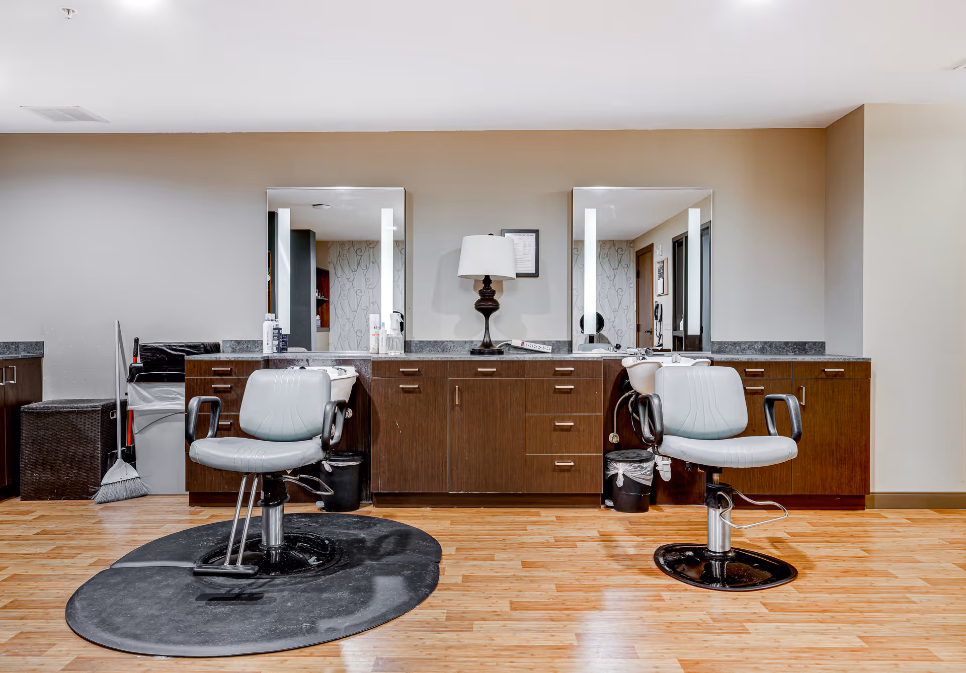 Interior view of a salon area with two gray salon chairs placed in front of a long wooden cabinet with two large mirrors. A lamp is positioned between the mirrors on the cabinet. The floor is wooden and there are cleaning tools like a broom and trash bins visible on the left side.