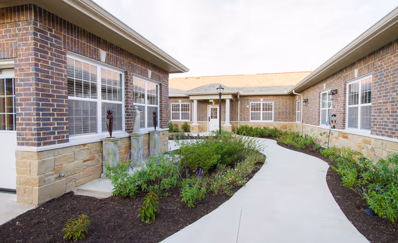 Outdoor courtyard area of Silverado Barton Springs Memory Care Community featuring a curved concrete walkway surrounded by landscaped garden beds with green shrubs and flowers. The building exterior is made of brick and stone with multiple windows and decorative statues near the entrance.