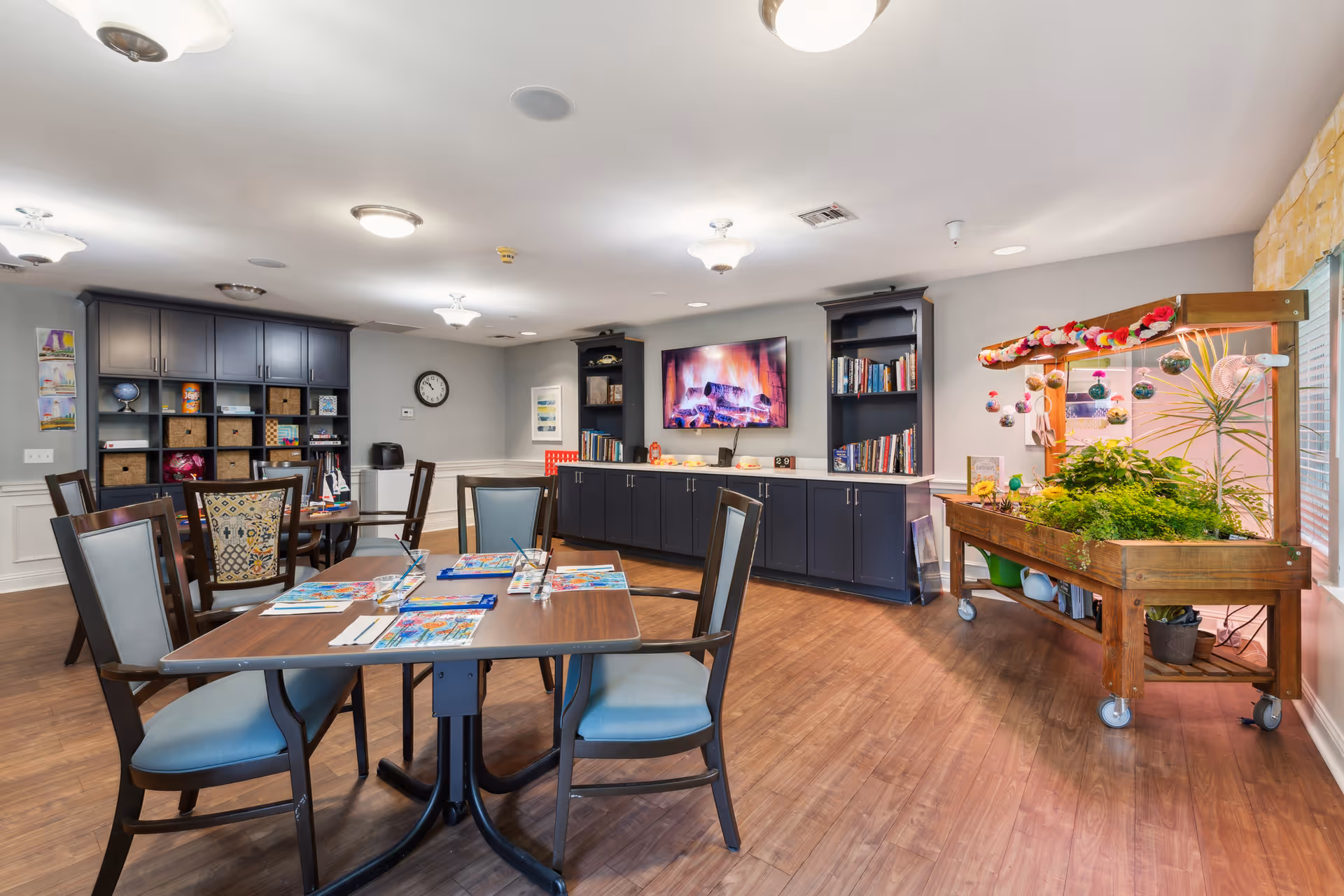 A bright and spacious common area with wooden flooring, several tables and chairs arranged for activities, a large cabinet with shelves and storage baskets, a wall-mounted TV displaying a fireplace scene, bookshelves filled with books, and a wooden plant stand with various green plants and hanging decorations near a window.