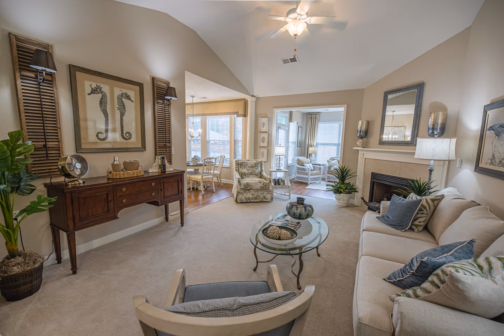 A cozy living room with a beige sofa adorned with blue and patterned pillows, a glass coffee table with decorative items, a fireplace with a mirror above it, and a wooden sideboard with seahorse artwork on the wall. The room opens into a dining area with a table and chairs near large windows letting in natural light.
