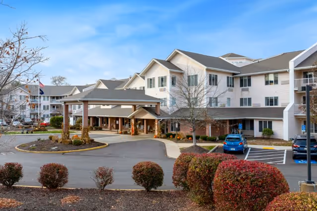 Front entrance of a multi-story senior living facility with a covered porte-cochere, parking area, and landscaped shrubs.