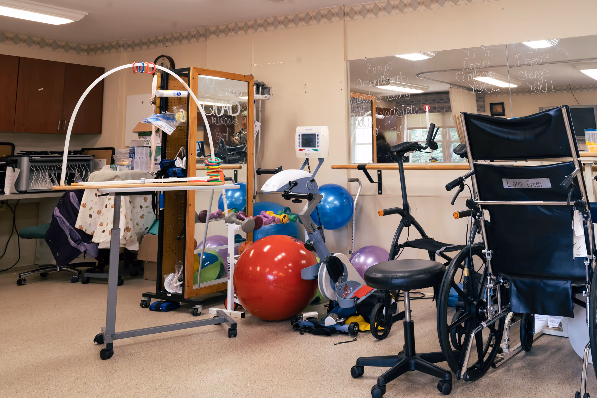 A therapy or rehabilitation room with exercise equipment including a stationary bike, large red and blue exercise balls, a wheelchair labeled 'Long Green', and various therapy tools. There is a mirror on the wall with motivational writing, and a desk with office supplies in the background.