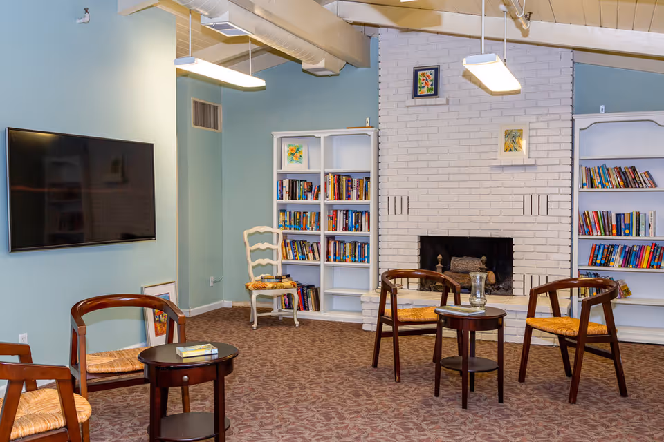 A cozy senior living common area with a white brick fireplace, two white bookshelves filled with books, four wooden chairs with woven seats arranged around two small round tables, a flat-screen TV mounted on a light blue wall, and framed artwork above the fireplace and on the bookshelves.