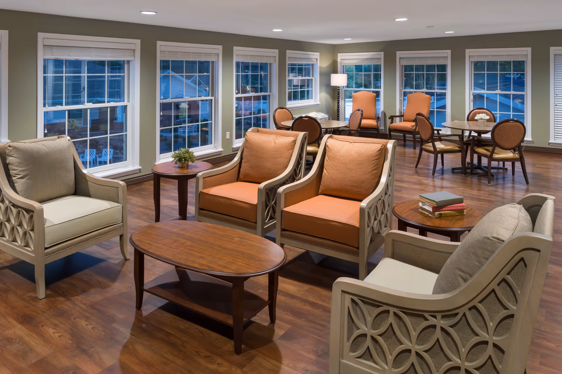 A spacious and well-lit common area with multiple armchairs and small round tables arranged on a wooden floor. Large windows line the walls, allowing natural light to fill the room. The seating includes cushioned chairs in beige and orange tones, with some tables holding books and a small plant. The room has a calm and inviting atmosphere.